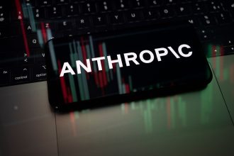 The logo of Claude artificial intelligence developed by Anthropic appears on a smartphone screen placed on a laptop computer displaying financial market curves.